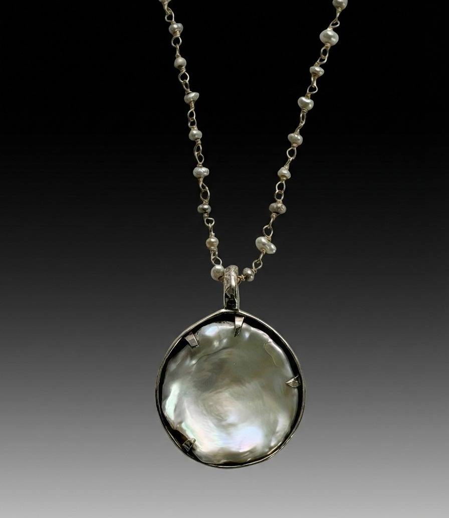 A round, iridescent mother of pearl pendant set in silver hangs from a delicate chain adorned with small pearls against a gradient gray background.
