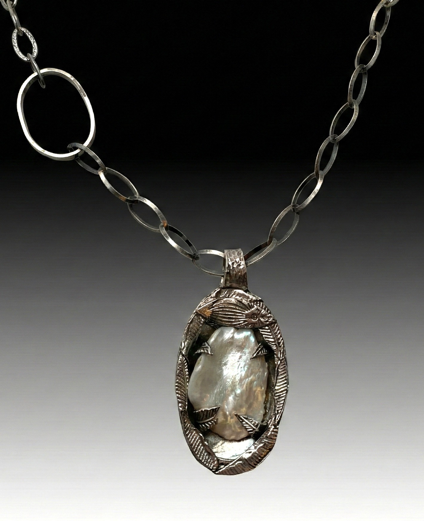 A silver necklace with an oval pendant featuring an iridescent, pearl-like stone set in a textured metal frame, on a dark gradient background.