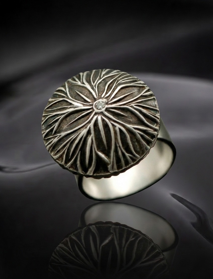 A round silver ring with a textured floral design and a small central diamond is showcased against a dark, reflective surface, exuding elegance.