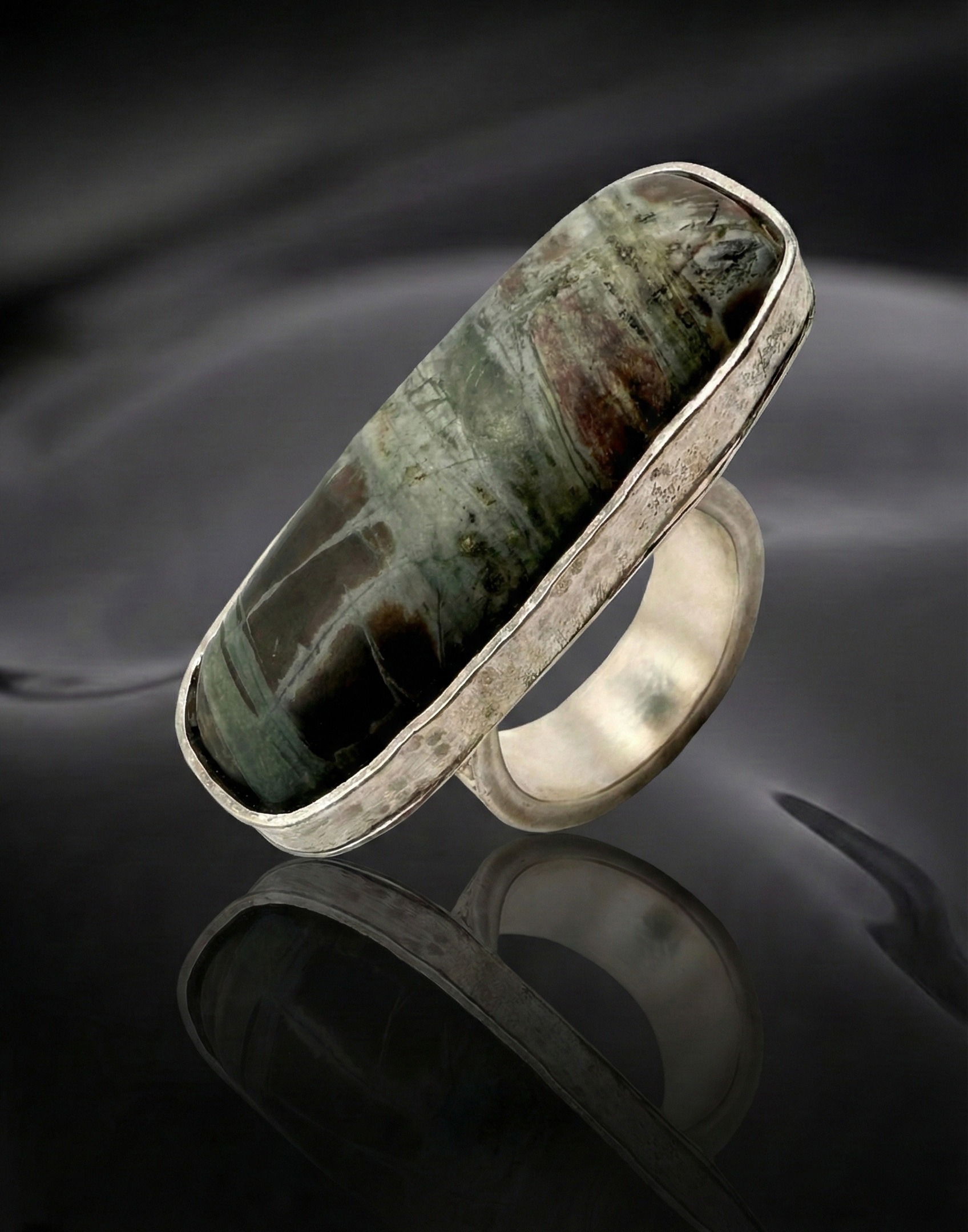 A silver ring with a large, elongated stone featuring dark green and brown hues. The background is a glossy black, reflecting the ring below.