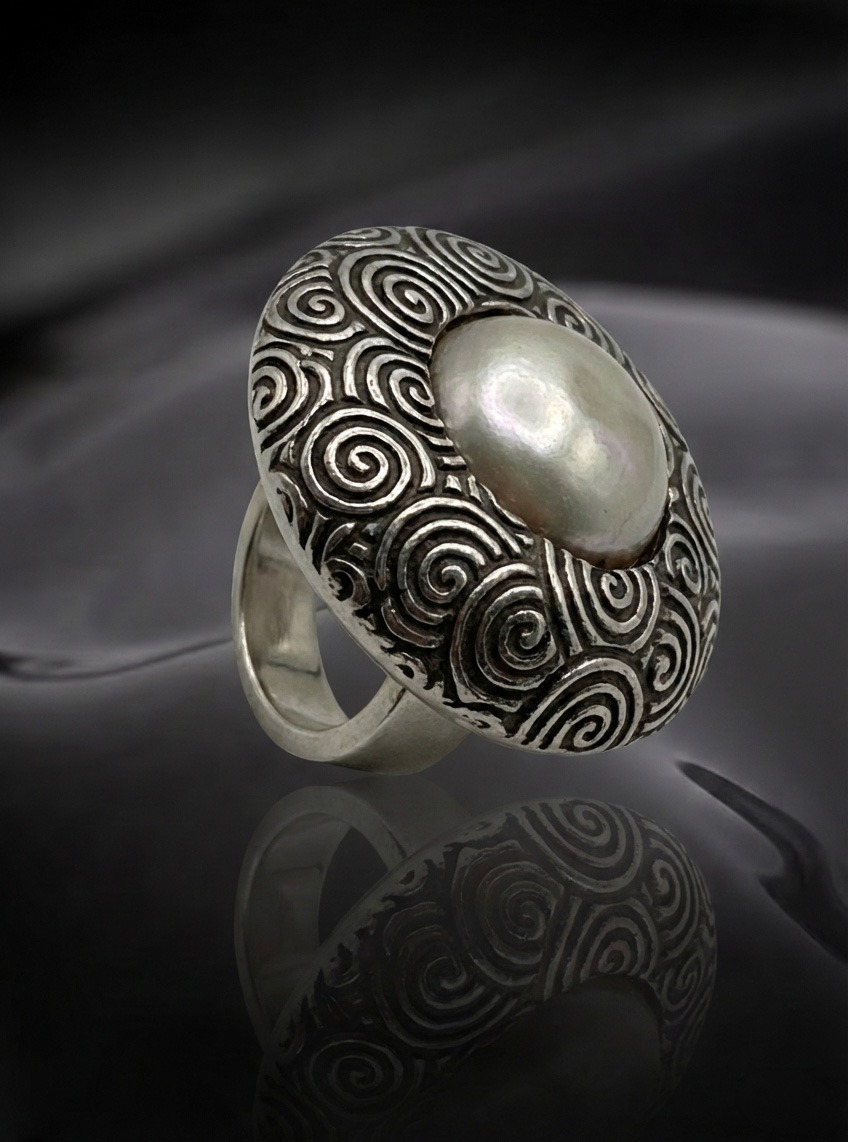 Silver ring adorned with a swirling patterned design, featuring a prominent pearl centerpiece. Reflective surface enhances its elegance.